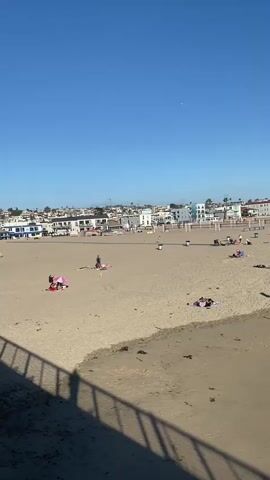 The video shows a beach scene with a clear blue sky and a sandy beach. In the foreground, a wooden pier with a railing casts a shadow on the sand. There are several people on the beach, some sitting under umbrellas or towels, and others walking or standing. In the background, there are buildings that appear to be houses or businesses, and palm trees. The beach is relatively empty, suggesting it is not a crowded day. There are some orange cones on the beach, possibly indicating a designated area or a temporary setup. The overall impression is a peaceful day at the beach.