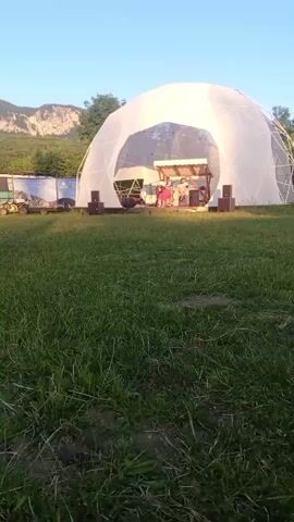 Two young girls are dancing in a grassy field in front of a large white geodesic dome. The dome appears to be a stage with a band performing inside. There are mountains in the background and a clear blue sky. A bus or RV is parked to the left of the dome, and some equipment is visible near the stage. The overall scene suggests an outdoor festival or event.