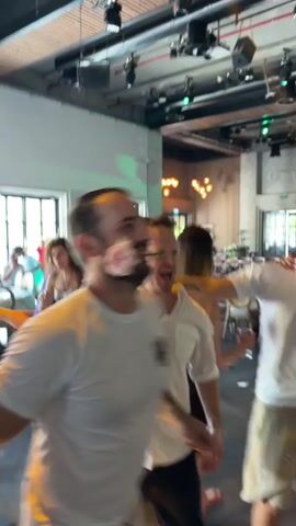 The video shows a group of people dancing at a party. The camera focuses on a man in a white t-shirt who is smiling and gesturing with his hands. He appears to be the center of attention. Other people are blurred in the background, some dancing and some standing. The environment appears to be an indoor venue with a ceiling with lights and exposed beams. There is a general atmosphere of celebration and joy.