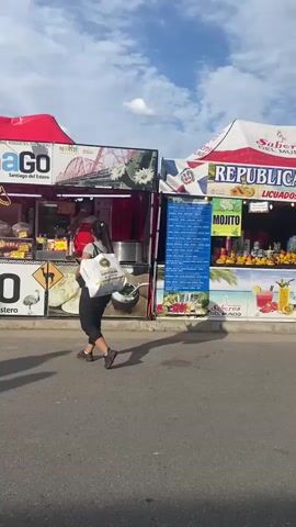The video shows a busy street scene at an outdoor event or fair.  There are multiple food vendor booths, each decorated with different national flags, including Argentina, Turkey and Ireland. People of all ages are walking around, browsing the booths, and talking.  Some appear to be making purchases, while others are just observing. The weather is sunny with some clouds in the sky. The setting is urban, with streetlights visible.