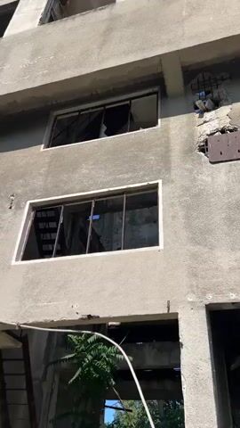 The video shows a dilapidated concrete building with broken windows. An external metal staircase, rusted and in disrepair, leads up to an opening in the building. Overgrown vegetation is visible both inside and outside the structure. The ground is covered in rubble and gravel.