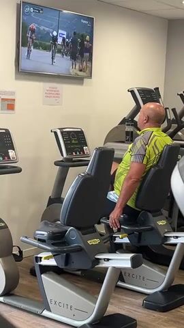 A man with a bald head and a green and yellow patterned shirt is sitting on a stationary exercise bike in a gym. He is looking up at a flat-screen television mounted on the wall, which is displaying a video of a cycling race. The man's hands are resting on the handlebars of the bike. The gym appears to be modern with other exercise machines visible in the background. There is a small sign with text on the wall below the television. The lighting in the gym is bright.