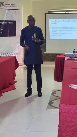 The video shows a presenter at a meeting or conference. He is a dark-skinned man wearing a dark blue long-sleeved shirt and black pants. He is holding a microphone and speaking to a group of people seated at a long table covered with a red tablecloth. The attendees are taking notes and listening to the presenter. A projector screen in the background displays a presentation with the title "How Divisive Narratives Are Built Locally." Banners are visible on either side of the screen, one reading "TETRA TECH International Development" and another "High-Level Dialogue on Divisive Narratives and Promoting Social Cohesion." The room appears to be a conference room with tiled floors. The overall setting suggests an academic or professional discussion on social issues.
