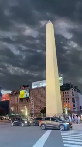 The video shows the Obelisco de Buenos Aires, illuminated at night, with dark, heavily clouded skies above. Street traffic, including cars and a motorcycle, moves in the foreground. Several buildings are visible around the obelisk, adorned with advertisements.