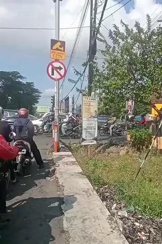 This is a street-level video taken on a sunny day with a clear sky. The camera is positioned on a sidewalk next to a road where traffic is moving. In the foreground, several motorcycles are visible. One motorcycle has a rider wearing a red jacket and a black helmet. Another motorcycle has a rider wearing a blue backpack and a red helmet. Behind these motorcycles, there is a white car, a white van, and several other motorcycles. A traffic light is visible on a pole, along with a sign indicating CCTV surveillance and another sign with a red circle and slash over a right-pointing arrow, signifying "no right turn." Trees and vegetation are present on the right side of the road. The road appears to be a multi-lane street.