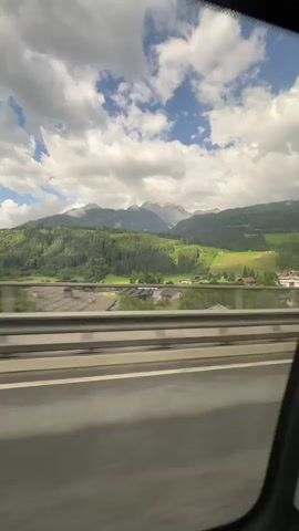 The media file shows a view from a moving vehicle, likely a car, traveling along a highway. The foreground is dominated by the vehicle's interior, with a blurred perspective of the road and a metal guardrail. Beyond the guardrail, a scenic landscape unfolds. Lush green hills covered in dense forests rise dramatically, forming the backdrop to a valley dotted with houses and other structures. In the distance, majestic mountains with snow-capped peaks pierce a sky filled with dramatic, puffy white clouds against a vibrant blue. The scene suggests a journey through a mountainous region, possibly the Austrian Alps, given the mention of Schlaming. The time of day appears to be daytime, with ample sunlight illuminating the landscape, although the presence of heavy clouds indicates a dynamic weather pattern. The overall impression is one of natural beauty and a sense of travel and exploration. No people or specific objects are clearly discernible as subjects, but the focus is on the expansive and picturesque scenery. There is no visible text in the media file.