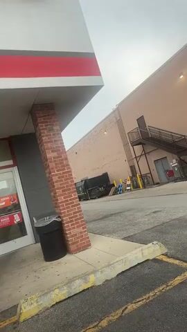 The video shows an exterior view of a building, possibly a commercial establishment. In the foreground, a brick column supports an overhang with red and white trim. To the left, a black trash can is visible. The background features a large, light-brown building with a metal staircase leading to an upper-level door. Below the staircase, another door is visible with the number 6481 above it. Several yellow bollards and a red shopping cart are also present. Additionally, there are large dumpster containers and stacked pallets. The sky is overcast.