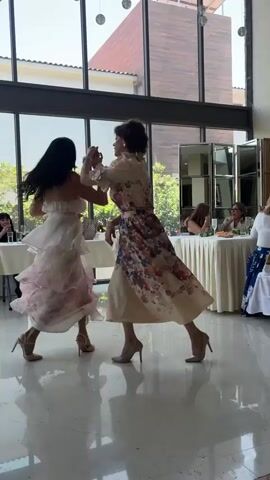 Two women are dancing at an indoor event, possibly a wedding reception or a formal party. They are both wearing elegant dresses and high heels. The woman on the left is wearing a strapless, tiered white dress with subtle pink floral accents. The woman on the right is in a cream-colored dress with voluminous puffed sleeves and a full skirt adorned with a vibrant floral print. They are holding hands and appear to be enjoying themselves, with smiles on their faces.

The setting is a well-lit indoor space with large windows that offer a view of greenery outside, suggesting it might be daytime. The floor is reflective, creating mirror images of the dancers. In the background, tables are set for a meal, covered with white tablecloths, and other guests are seated and conversing. Mirrors are strategically placed on one wall, adding to the spacious feel of the room. The overall atmosphere seems celebratory and joyful. There is no visible text in the media file.
