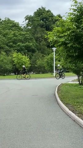 Two individuals are cycling on a paved path lined with trees and grass. The person on the left is riding a yellow bicycle and wearing a white helmet and black clothing. The person on the right is riding a black bicycle and wearing a green and black cycling suit with a white helmet. In the background, a white lamppost is visible. Two additional individuals, one wearing a beige jacket and the other a red jacket, are walking on the path in the lower left portion of the frame. The sky is overcast.