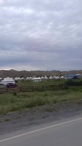 The video shows a landscape with rolling hills in the background under a cloudy sky. In the middle ground, there is a cluster of traditional round dwellings, possibly yurts, arranged in a semi-circle. Some of these structures are white, while others appear to be made of lighter material. A dark blue building with a dark roof is situated among the yurts. A wooden fence runs horizontally across the middle of the frame, separating the dwellings from a grassy field in the foreground. To the right, an arched gateway with decorative elements is visible, and flags are placed along the top of the gateway. The foreground consists of a paved road surface. The camera appears to be moving, suggesting the footage was captured from a vehicle.