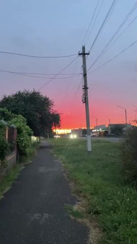 The video shows a rural road at dusk. The sky is filled with a vibrant pink and orange sunset, transitioning to a pale blue overhead. A sidewalk runs along the left side of the road, bordered by lush green grass and trees. A power pole with numerous wires stands prominently in the foreground, to the right of the sidewalk. In the distance, there are buildings, streetlights, and a few cars driving on the road, their headlights illuminated. The overall atmosphere is peaceful and serene, capturing the beauty of a rural sunset.