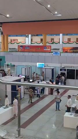 The video shows the interior of a building, likely a train station or airport, with an upper level and a main floor. On the main floor, there is a security checkpoint with metal detectors and X-ray machines. People are lining up to go through security. Some security personnel are visible, wearing uniforms. There are also counters and desks with people working at them. The building appears to be well-lit and decorated with flags and banners. The overall atmosphere seems to be one of orderly movement and security screening.