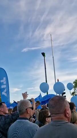 A large crowd of people are gathered outdoors, looking up at the sky. Many of them are holding blue balloons and blue, fluffy umbrellas. In the background, there are modern brick buildings with large windows and trees. The sky is blue with wispy white clouds, suggesting it is daytime. The overall atmosphere appears festive.