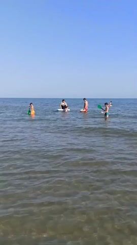 The video shows a group of people in the ocean on a sunny day. The water is clear and calm, with gentle waves. The sky is a clear blue. In the foreground, the sandy bottom is visible through the water. In the middle ground, there are several people in the water. From left to right, a person in an orange swimsuit stands in the water, holding a green paddle. Next, a person in a black swimsuit is sitting on a white floatation device. To their right, a person in red swim trunks stands in the water, and a child is partially submerged next to them. Further to the right, a person in blue swim trunks is standing in the water, holding a green paddle and a white object. In the background, two more people are visible in the distance, one on a floatation device and another standing in the water. The people appear to be playing or relaxing in the ocean.
