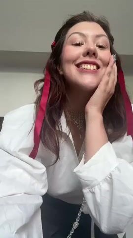 A young woman with dark hair adorned with red ribbons smiles and poses for a video. She wears a white wrap shirt, a silver necklace, and a grey pleated skirt. A small black purse with a silver strap hangs across her body. She touches her face and then moves her hands to her hair, smiling and dancing. She turns her back to the camera and then faces forward again, her hands moving expressively as she continues to smile and dance. The background is a plain wall.