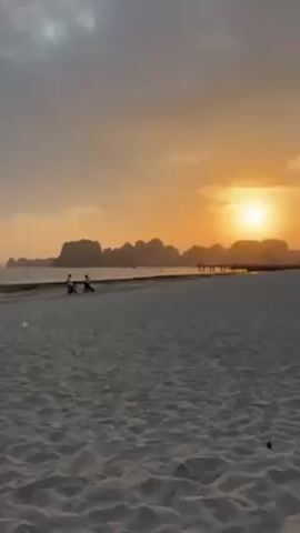 The video shows a sandy beach at sunset. The sky is a mix of orange, yellow, and gray.  The sun is setting behind a row of mountainous islands on the horizon. A few people are visible on the beach in the distance, and there's a pier or walkway extending into the water. Palm trees line the right side of the beach. The sand is light-colored and smooth, with footprints visible.  The camera appears to be panning slightly to the right.