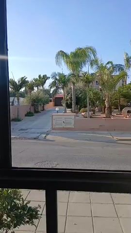 The video shows a street view from a window. On the left side of the street, there is a brown-colored residential complex with tall green palm trees. The complex has a long driveway leading to a garage. On the right side of the street, there is another building with a landscaped front yard filled with various plants and palm trees. The sky is clear and blue, indicating a bright day. The camera pans slightly to the left and right, capturing different angles of the street and buildings. There are no people visible in the video.
