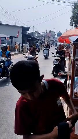 The video shows a street scene. In the foreground, a young male wearing a red t-shirt is partially visible, looking downwards. He is in silhouette. Behind him, a street runs through a town or village. Several motorcycles are visible on the road. One motorcycle in the mid-ground carries two passengers, both wearing helmets and facing forward. Another motorcycle further back also carries two people, one driving and one as a passenger. In the background, buildings line the street, and a dome-shaped structure is visible. The sky is bright and appears to be daytime. A food cart with an orange umbrella is present on the right side of the frame.