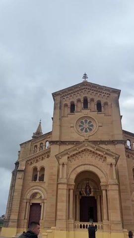 The video shows a large, tan-colored church with a cross on top, shot from a low angle. The sky is overcast and cloudy. The camera pans to the right, revealing an open plaza with a mosaic mural along the back wall. People are walking around the plaza. A distant hilltop with a white structure on top is visible.