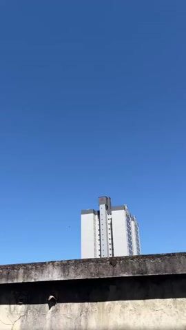 A concrete wall extends horizontally across the foreground of the video. Behind the wall, a tall, white apartment building with a gray section on its side rises against a clear blue sky. The building has numerous windows visible. A small, dark object, possibly a bird, is in flight in the sky to the left of the building. The camera is static.