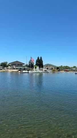 The video shows a wide shot of a lake on a clear, sunny day. The water is calm with gentle ripples, and the sunlight reflects off its surface, revealing the sandy bottom beneath. On the shore, there is a building with a tan-colored roof and several windows, possibly a restaurant or clubhouse. An American flag flies prominently from a flagpole in front of the building. Several sailboats are moored near the shore, with their masts visible against the bright blue sky. Another, larger building with a green roof and white walls is visible further down the shoreline, along with a line of smaller structures. Trees and some shrubbery dot the landscape. The scene suggests a recreational area, likely a marina or a lake resort, and the weather indicates it's midday or afternoon. The overall mood is peaceful and serene, characteristic of a calm day at the water. No visible text is present in the video.
