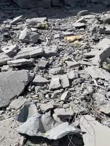The video shows the ruins of a building under a bright sky. Debris and rubble cover the ground. The building’s remaining structure consists of cracked and broken concrete pillars and ceilings. At one point, a person dressed in white walks through one of the dilapidated doorways into the building.
