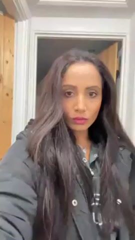 A woman with long, dark hair, wearing a black puffer jacket and a grey shirt with white writing, stands in what appears to be a hallway. The background includes a white door frame and a partially visible wooden door on the right. The woman is looking directly at the camera and puckering her lips.