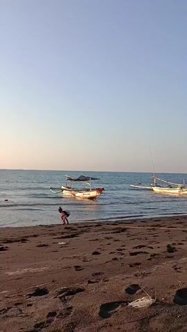 A vertical, eye-level shot captures a beach scene during daylight. The sand in the foreground is dark brown and littered with footprints and some white plastic debris. The water is calm and a light blue, meeting the shore with gentle waves. Two traditional fishing boats are moored in the water. The boat on the left is white with a reddish-orange trim and a dark canopy. The boat on the right is entirely white and has a tall mast. A person in red clothing is crouched near the waterline in the middle left of the frame. The sky is a clear, pale blue, transitioning to a lighter hue near the horizon.