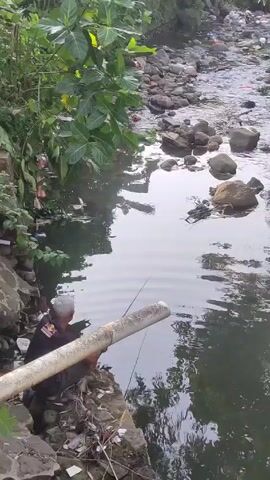 A person is fishing in a small stream. The person is sitting on a rocky bank, next to a large pipe. The stream is surrounded by vegetation and rocks, and there is some trash visible in the water and on the banks. The water appears relatively calm.