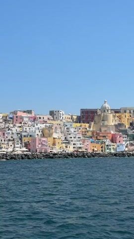 The video shows the colorful buildings of Procida, Italy, from the water. The buildings are a mix of colors, including pink, yellow, white, and red. The water is a dark blue, and the sky is clear. There are a few boats in the water near the rocks, and some birds fly across the screen. The camera is moving slightly, and the music is playing.