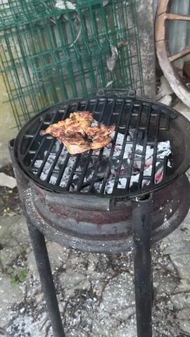 The video shows chicken being grilled on a small, round, charcoal grill. The grill is old and weathered, with ash visible in the bottom. The chicken has grill marks and is steaming slightly. The background includes a green wire fence, some concrete, and a portion of a wooden wagon wheel. No people are visible.