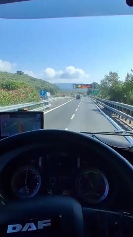 The video is filmed from the perspective of a driver inside a DAF truck, looking forward through the windshield. The truck is driving on a highway under a clear blue sky with scattered white clouds. A digital sign is visible ahead, displaying traffic information in Italian, indicating a 1 km traffic jam near Finale Ligure. The highway has multiple lanes separated by barriers. There are trees and hills on the left side of the road and barriers on the right. Other vehicles, including cars and a truck, are visible on the highway. A GPS device is mounted on the dashboard to the left. The steering wheel of the truck is prominent in the foreground. The scene depicts a sunny day on an Italian highway.