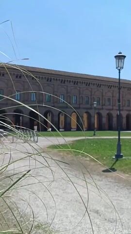 The video shows a long, brick building with a series of arches in front of it. There is a green lawn in front of the building, and a lamp post is standing on the lawn. A small gravel path is in front of the lawn, and some long grasses are blurred in the foreground. The sky is blue and clear.