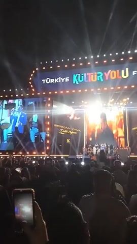 A crowd of people are gathered at night to watch a live music performance on a large outdoor stage. The stage has a large screen behind the band displaying images of the performing artist, Serkan Kaya, along with the album title "Dedikoducu Meyhane". The stage design also includes the words "Türkiye Kültür Yolu Festivali" and "Kültür ve Turizm Bakanlığı" (Ministry of Culture and Tourism) illuminated in lights, suggesting the event is part of a government-sponsored cultural festival. Many people in the audience are holding up their phones, presumably to take pictures or record videos of the performance. The lighting is bright and dynamic, with spotlights shining on the stage.