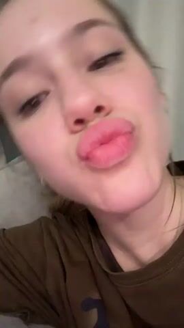 This is a close-up video of a young Caucasian woman with brown hair. She is wearing makeup, including mascara and pink lipstick, and has her head tilted to the side, resting on a brown surface. The lighting appears to be indoor, and a blurry blue and grey background is visible. She makes a kissing gesture and then smiles slightly. The video consists of several short clips that create a slideshow effect.