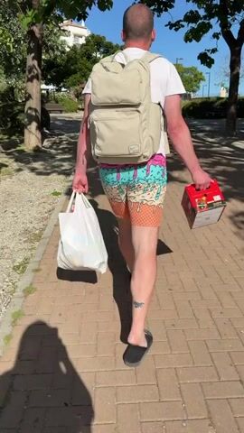 A man is walking down a brick sidewalk. He is wearing a white T-shirt, ice cream cone-patterned shorts, and black flip-flops. He has a beige backpack on his back and is carrying a white plastic shopping bag in his left hand and a red box of beer in his right hand. The sidewalk is lined with trees that provide shade. In the background, there is a bus, a park, and other pedestrians.