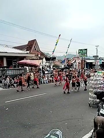 A group of people dressed in red and white attire are participating in a parade on a paved road. They appear to be performing a ritualistic dance or procession, holding long sticks and moving in unison. The parade is taking place in a village or town setting, with buildings and shops lining the street. Several motorcycles are parked on the side of the road, and spectators can be seen watching the event. The sky is overcast, suggesting a cloudy day. The overall atmosphere seems festive and communal, indicating a local celebration or cultural event.