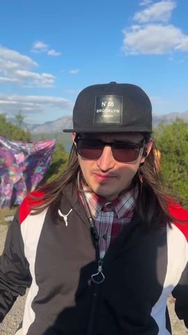 A man wearing a black baseball cap with "N'86 BROOKLYN TWENTY" on the front, sunglasses, a black, white, and red Puma jacket over a plaid shirt, black pants, and grey sneakers is standing on a dirt road. He has long brown hair. He is talking, and he is gesturing with his hands. Behind him are various objects, including a colorful piece of fabric and an orange stuffed animal. In the background is a mountain range and a body of water. The sky is blue with some clouds.