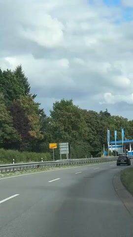 The video shows a view from inside a vehicle driving on a road. The road curves to the right. On the left side of the road, there is a guardrail, trees, and a yellow sign with black text. On the right side of the road, there is a black car driving ahead and a gas station with a blue roof and white pillars. The gas station has several pumps and signage. The sky is cloudy with patches of blue. The overall environment appears to be a roadside scene in a suburban or rural area.