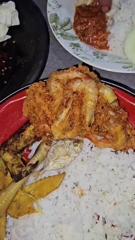 A high-angle, close-up shot shows two plates of food and a person's hands. On the top plate, white rice is topped with a scoop of red chili paste, a slice of cucumber, and grilled chicken. A person's hand reaches into the frame and appears to be mixing the chili paste with the rice. On the bottom plate, white rice is covered with fried shrimp and small fried fish. A black stone mortar filled with a dark sauce, chopped red onions, chili seeds, a lime wedge, and shredded cabbage is visible in the foreground. The background is a gray, textured surface, and a portion of a person's leg and foot is visible. The lighting is dim, suggesting an indoor or evening setting.