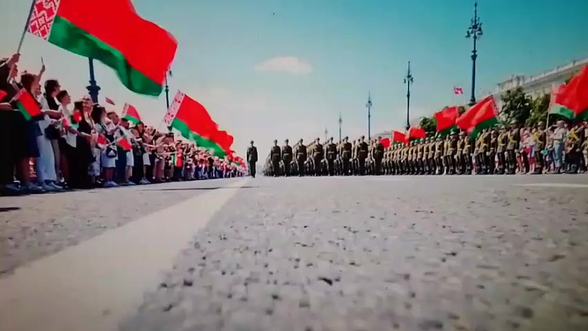 The video shows a low-angle shot of a street procession. On the left side of the street, a crowd of people are waving Belarusian flags. On the right side of the street, a line of soldiers in uniform are marching. A lone soldier walks in the middle of the street between the crowd and the marching soldiers. The sky is blue with a few clouds. The scene appears to be a formal event, possibly a parade or military ceremony.