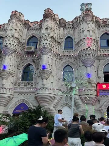 The video shows a crowd of people gathered in front of a large, ornate building that resembles a castle. The building is adorned with lights and decorative elements, including what appears to be a light display resembling a dandelion or fireworks. People are standing, looking at the building, and some are holding fans. There are trees and bushes in the foreground. The sky is visible in the background. A red banner with Chinese characters is displayed on the building. The overall atmosphere appears to be one of a public gathering or event.