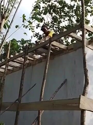 A low-angle, medium shot shows a construction worker on the roof of a partially built structure. The structure has white walls and a wooden frame for the roof. The worker, wearing a yellow shirt and a hat, is kneeling and appears to be working on the roof framing. He is positioned towards the right side of the frame. The environment is outdoors with trees visible in the background. The sky is overcast. The overall scene suggests ongoing construction work in a natural setting.