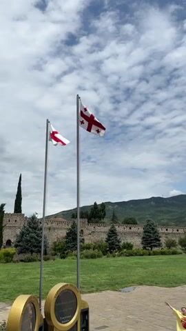 The video opens with a shot of two Georgian flags flying against a blue sky with white clouds. In the background, there is a stone wall with trees and a distant green mountain. A person in a yellow shirt with arms outstretched is visible in the lower right corner of the frame. The camera then pans to show a group of people walking on a paved area in front of the wall, with benches and green grass. The scene shifts to focus on a large, ancient stone church with a conical roof and a cross on top. People are visible near the entrance of the church. The camera continues to pan, showing more of the church's architecture and the surrounding landscape, with the mountains still visible in the background. The sky remains cloudy throughout the video.