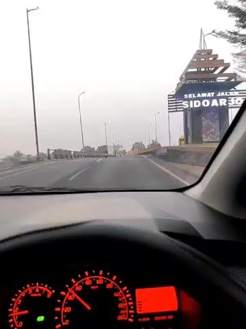 The video is filmed from inside a car, looking forward through the windshield. The car is driving on a highway overpass. The speedometer, which is red and digital, is visible in the lower left corner. The car appears to be traveling at a moderate speed. The highway is paved and has yellow and white lane markings. On either side of the highway are railings and streetlights. In the distance, several motorcycles can be seen on the road. The sky is overcast and a light gray. There are buildings visible in the background, including what appears to be a mosque. The overall scene suggests a cloudy day and a typical drive on an elevated highway.