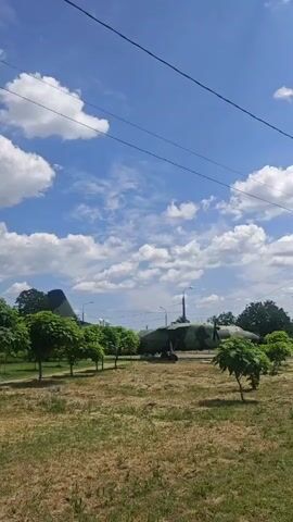 The video pans across a camouflage painted airplane parked on grass. The plane has a propeller on one side and a tail with a red star. There are trees in the foreground and a blue sky with clouds in the background.