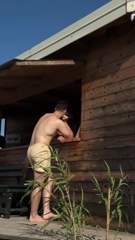 A man is at a small wooden shack. He is shirtless, wearing light-colored shorts. He is leaning on the sill of the shack's window, talking to someone inside. The shack has a sign that says "Staff Only". There is a security camera mounted on the side of the shack. There are tall green plants in front of the shack. A sign is seen in the background. The sky is blue.