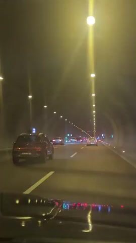 The video shows the view from a car driving through a tunnel. Several other cars are visible in the distance, illuminated by the tunnel lights. The tunnel is brightly lit with rows of overhead lights. The road is marked with white lines, and signs are visible on the sides of the tunnel. The car's dashboard is partially visible in the foreground.