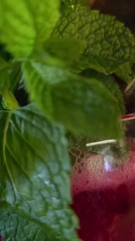 The media file shows a close-up of a vibrant, magenta-colored beverage in a clear glass goblet. The drink is garnished with fresh green mint leaves and has a frothy top. Two dark glass bottle necks are stuck into the drink, leaning against the mint. Colorful straws are also visible peeking out from behind the garnishes. The goblet sits on a light-colored wooden surface, and the background is blurred, showing hints of pink and copper tones, as well as some greenery. The lighting suggests it could be daytime, possibly indoors in a restaurant or bar setting, given the presentation of the drink. The overall impression is one of a refreshing and artfully prepared cocktail. There is no visible text in the media file.
