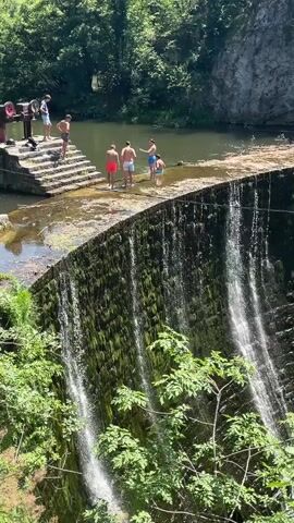 In Uzice, Serbia, a group of young people are enjoying a summer day by a waterfall. The scene is set by a curved dam with water cascading down its moss-covered face, creating a misty curtain. Lush green foliage, including trees and leafy branches, surrounds the waterfall, adding to the natural beauty of the location.

Atop the dam, several individuals, appearing to be teenagers and young adults, are gathered. Some are standing near the edge, looking down at the water, while others are wading in the shallow area. One person in a red swimsuit is seen bending down, possibly preparing to jump or splash. The overall atmosphere appears to be one of youthful exuberance and enjoyment of outdoor recreation.

In the background, beyond the dam, a calm body of water is visible, bordered by a rocky cliff face and dense green trees. There are also some man-made structures on the left side of the frame, including concrete stairs leading up to a platform with what looks like old machinery, possibly part of the dam's original infrastructure. The sunlight suggests it's a bright, clear day, ideal for swimming and spending time outdoors. The individuals' postures and proximity to the water convey a sense of relaxation and fun.